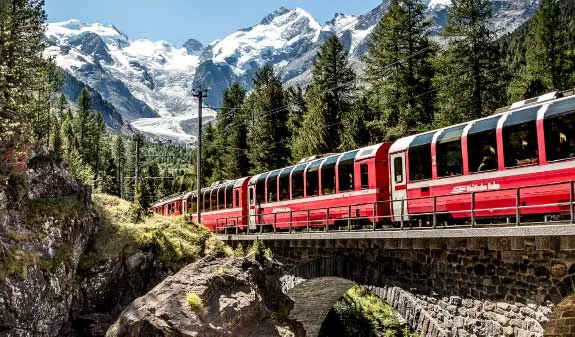 Roter Zug fährt durch bergige Landschaft mit schneebedeckten Gipfeln und grünen Nadelbäumen.