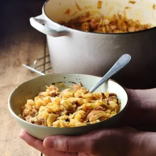 Sauerkraut-Eintopf in grüner Schüssel mit Löffel, von Händen gehalten, und grauem Topf mit Eintopf im Hintergrund.