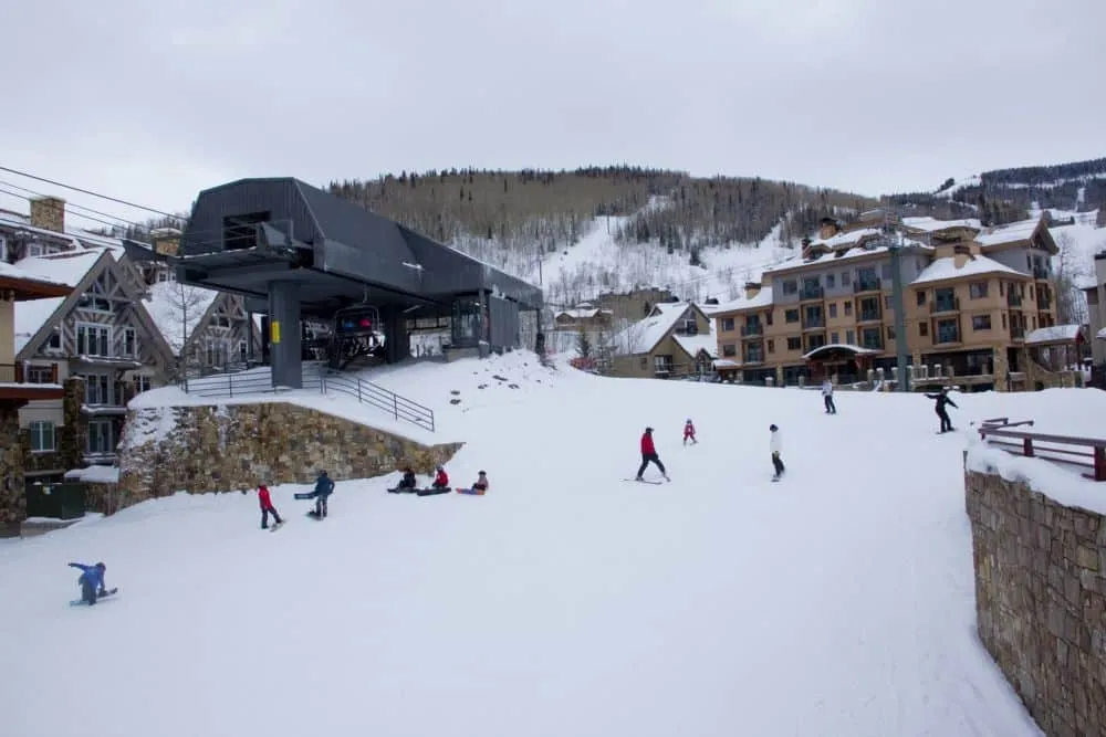 Skifahrer und Snowboarder gleiten in der Nähe eines Skilifts auf verschneiten Pisten in Colorado, umgeben von Hütten und Bergen unter einem bewölkten Himmel.