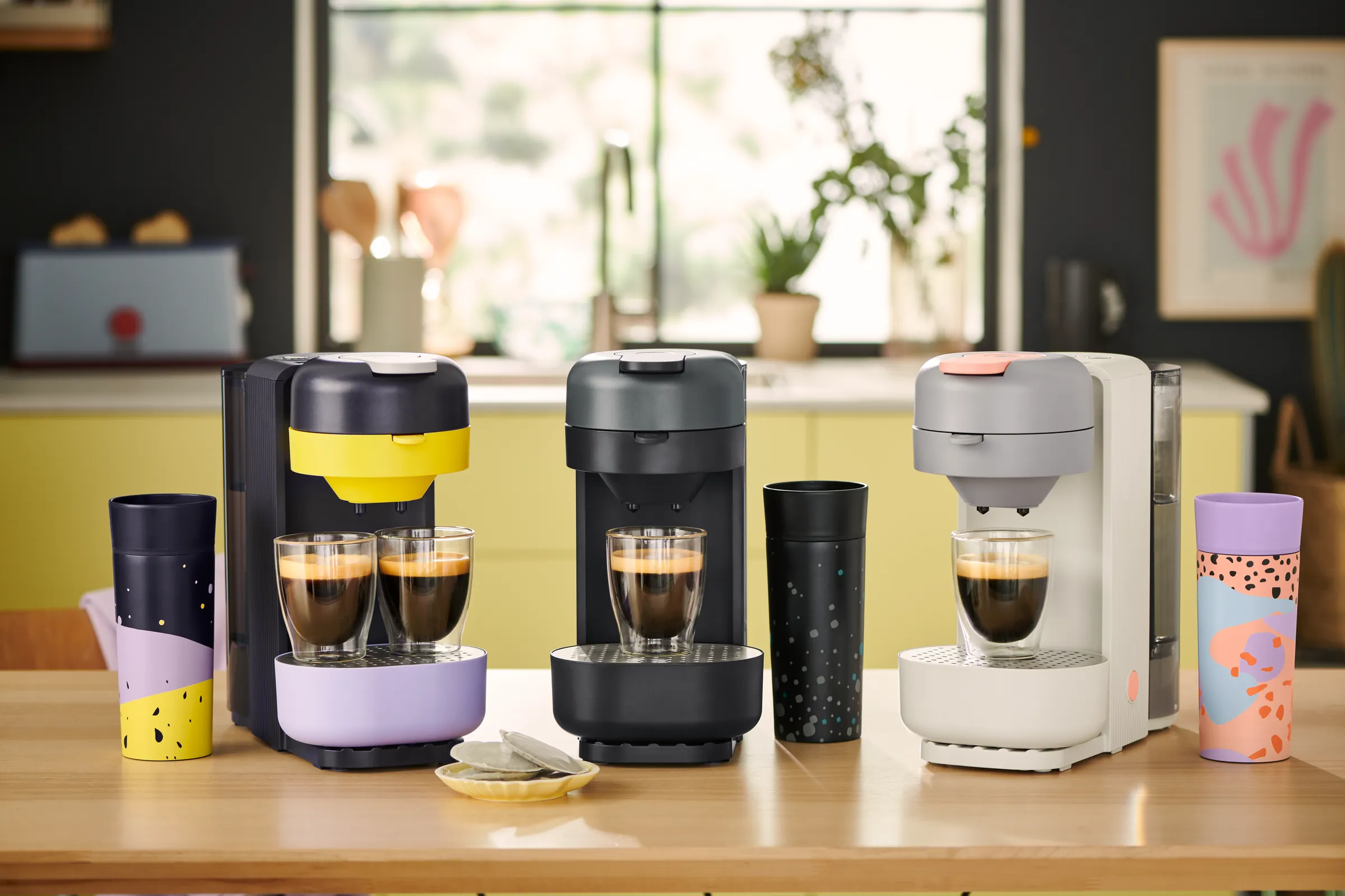 Sortiment der Tchibo CALL ME PAD Kaffeepadmaschine mit Pads, To-Go-Bechern und Milchaufschäumer in verschiedenen Farben.