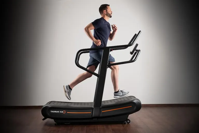 Sportler läuft auf dem Taurus Run-X Curved Treadmill. Das Laufband steht auf einem dunklen Holzfußboden vor einer weißen Wand.