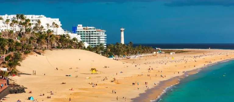 Strand von Morro del Jable auf Fuerteventura mit goldenem Sand und türkisblauem Meer. Im Hintergrund stehen weisse Gebäude und ein Leuchtturm, Palmen sind auf der linken Bildseite zu sehen.