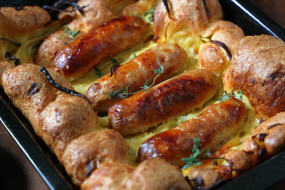 Traditionelles englisches &quot;Toad in the Hole&quot; mit Würstchen in Yorkshire-Pudding-Teig, umgeben von dunkler Bratensoße und frischer Petersilie.