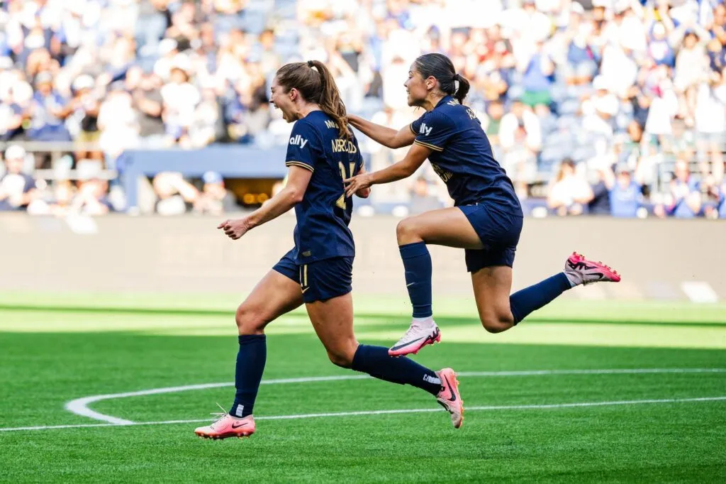 Two soccer players in navy blue uniforms celebrate on the field in a display of Seattle sports spirit. One player is running while the other jumps with excitement, extending an arm towards her teammate. The background shows a blurred crowd in a stadium.