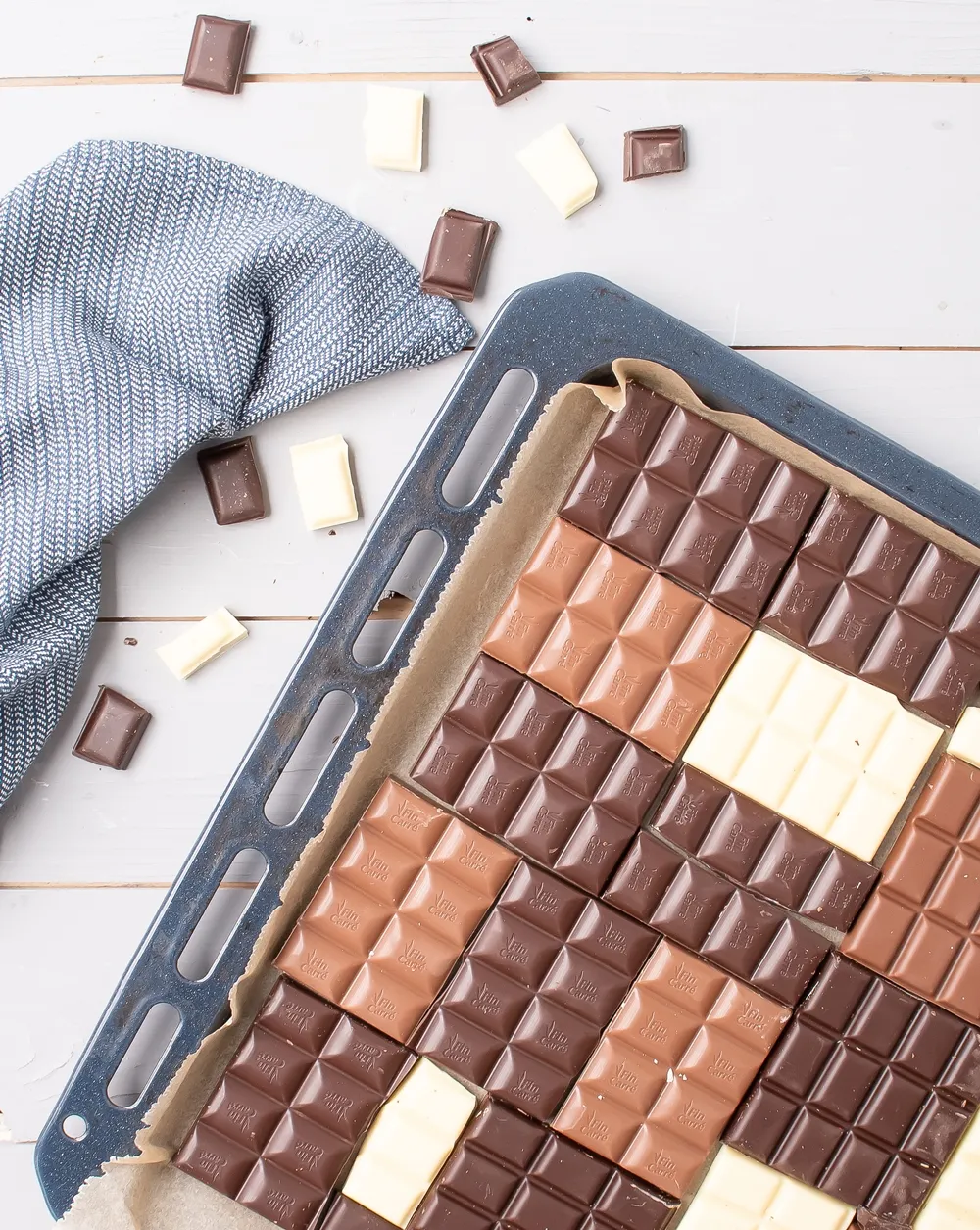 Verschiedene Schokoladentafeln (Zartbitter, Vollmilch, Weiß) kunstvoll auf einem mit Backpapier ausgelegten Blech angeordnet, bereit um Bruchschokolade selber zu machen.