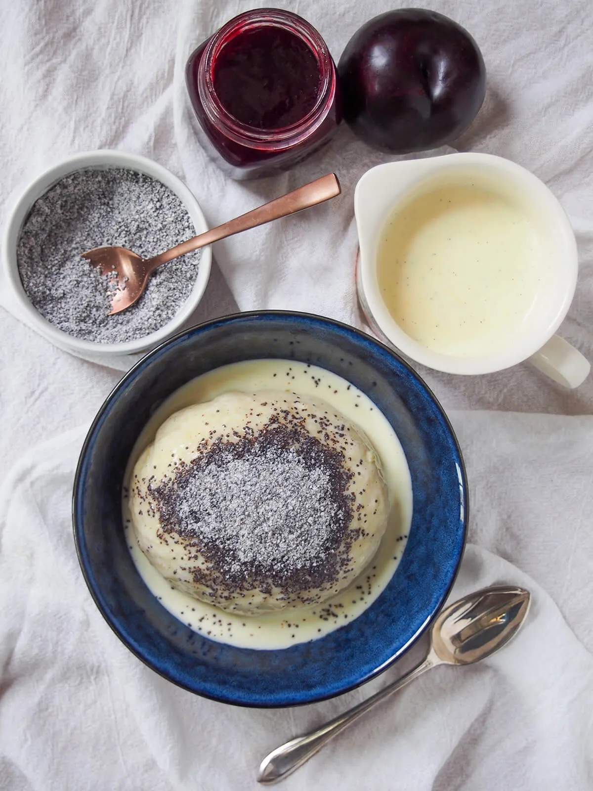 Vogelperspektive einer Schüssel mit Germknödeln, mit zusätzlicher Vanillesauce, gesüßtem Mohn und Pflaumenmarmelade über der Schüssel.