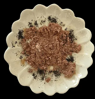 Weiße, blütenförmige Schale mit braunen, krümeligen Futtermischung und Zahnputzkohle auf der Oberfläche.