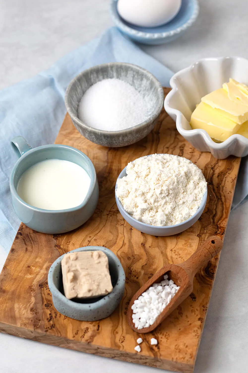 Zutaten für die Osternester – Mehl, Zucker, Butter, Eier und Milch in kleinen Schalen auf einem rustikalen Holzbrett angerichtet