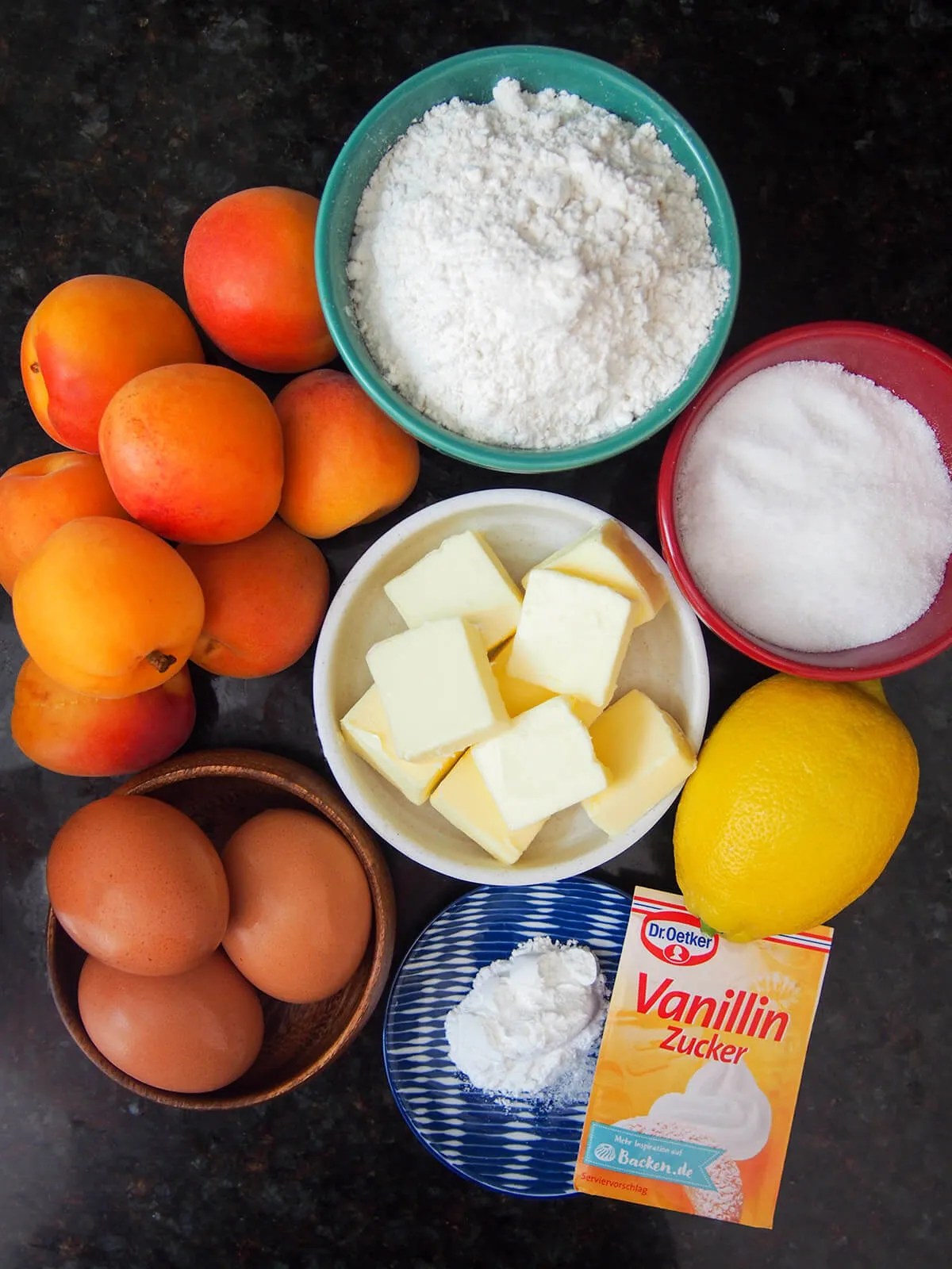 Zutaten für Marillenkuchen: Schüsseln mit Mehl, Butter, Zucker, Eier, daneben frische Aprikosen und eine Zitrone