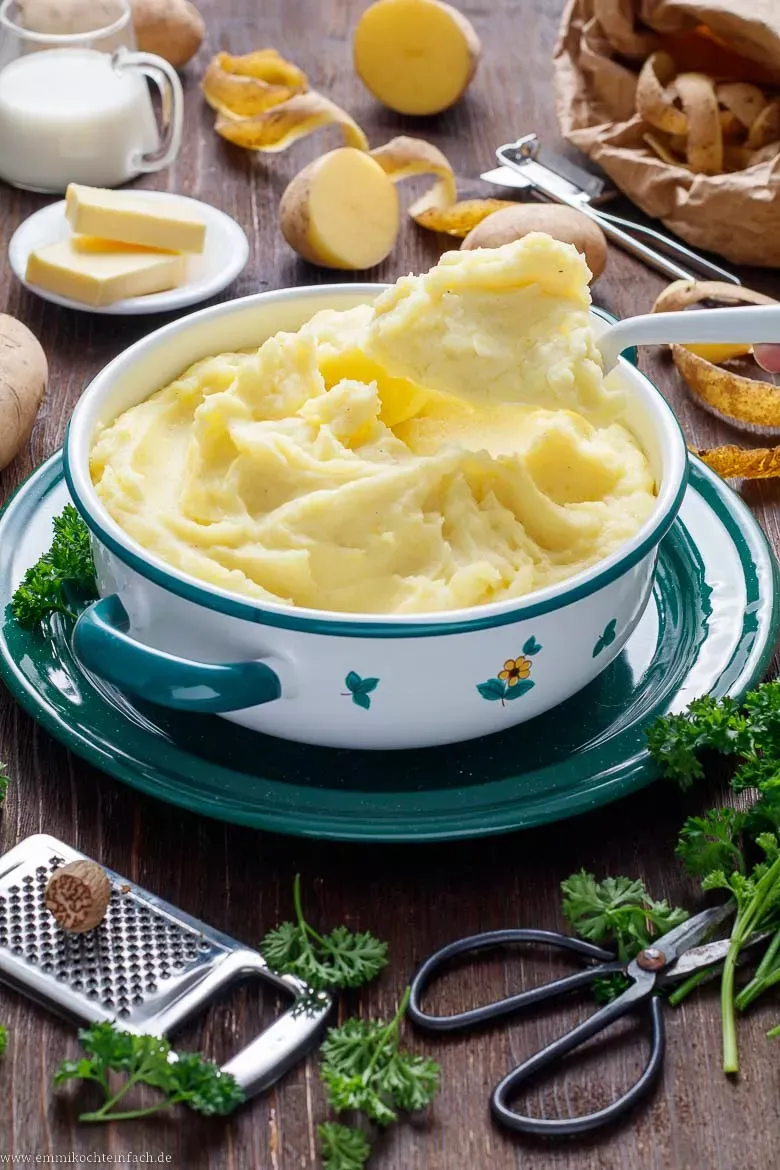 Zwei große Löffel schöpfen cremigen Kartoffelbrei aus einer weißen Schale, garniert mit frischer Petersilie und Muskatnuss