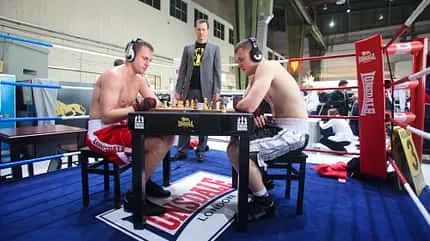 Zwei Männer in Boxshorts sitzen in einem Boxring an einem Schachbrett.