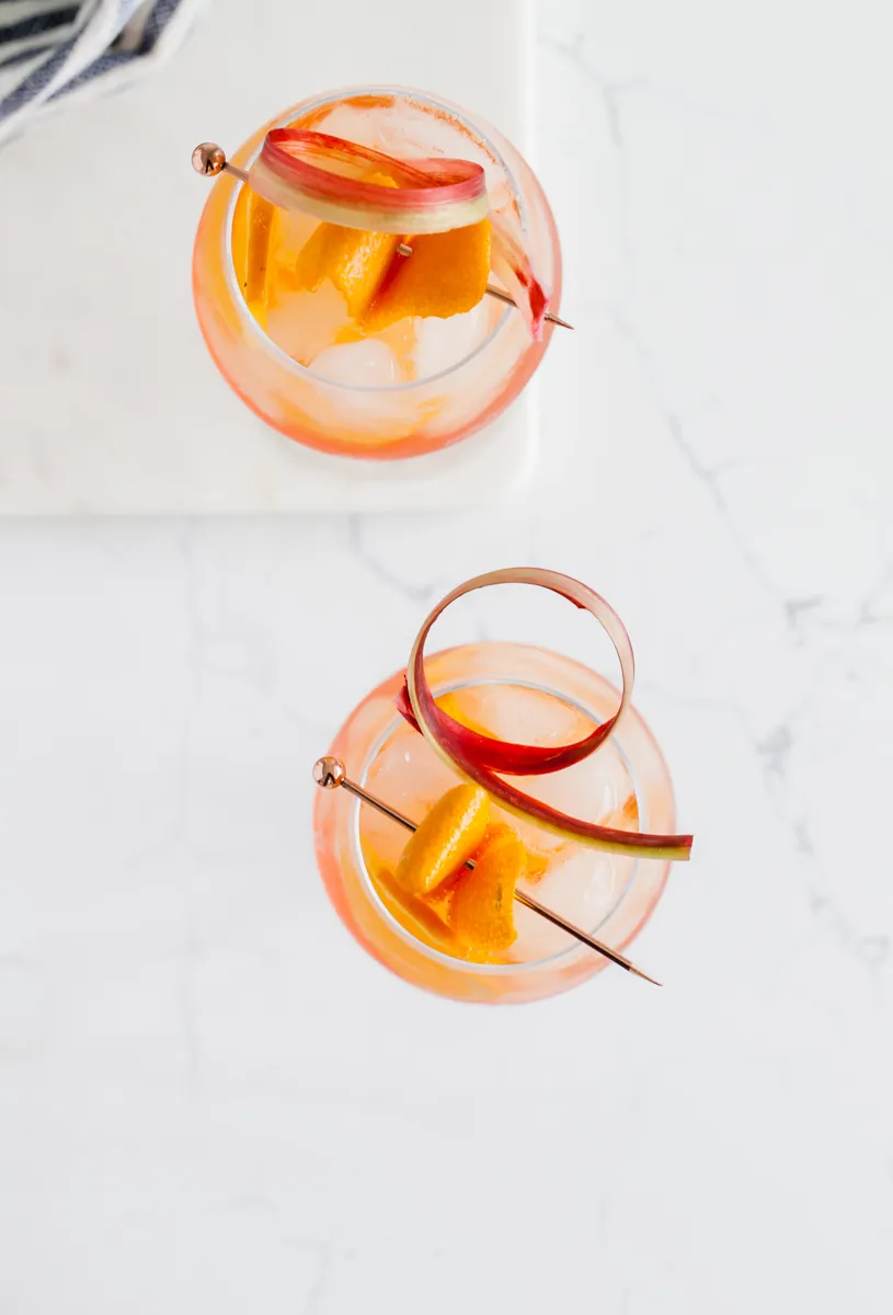 Zwei Rhabarber Aperol Spritz mit Eis, Orangenzeste und Rhabarberdeko vor einem verschwommenen Hintergrund.