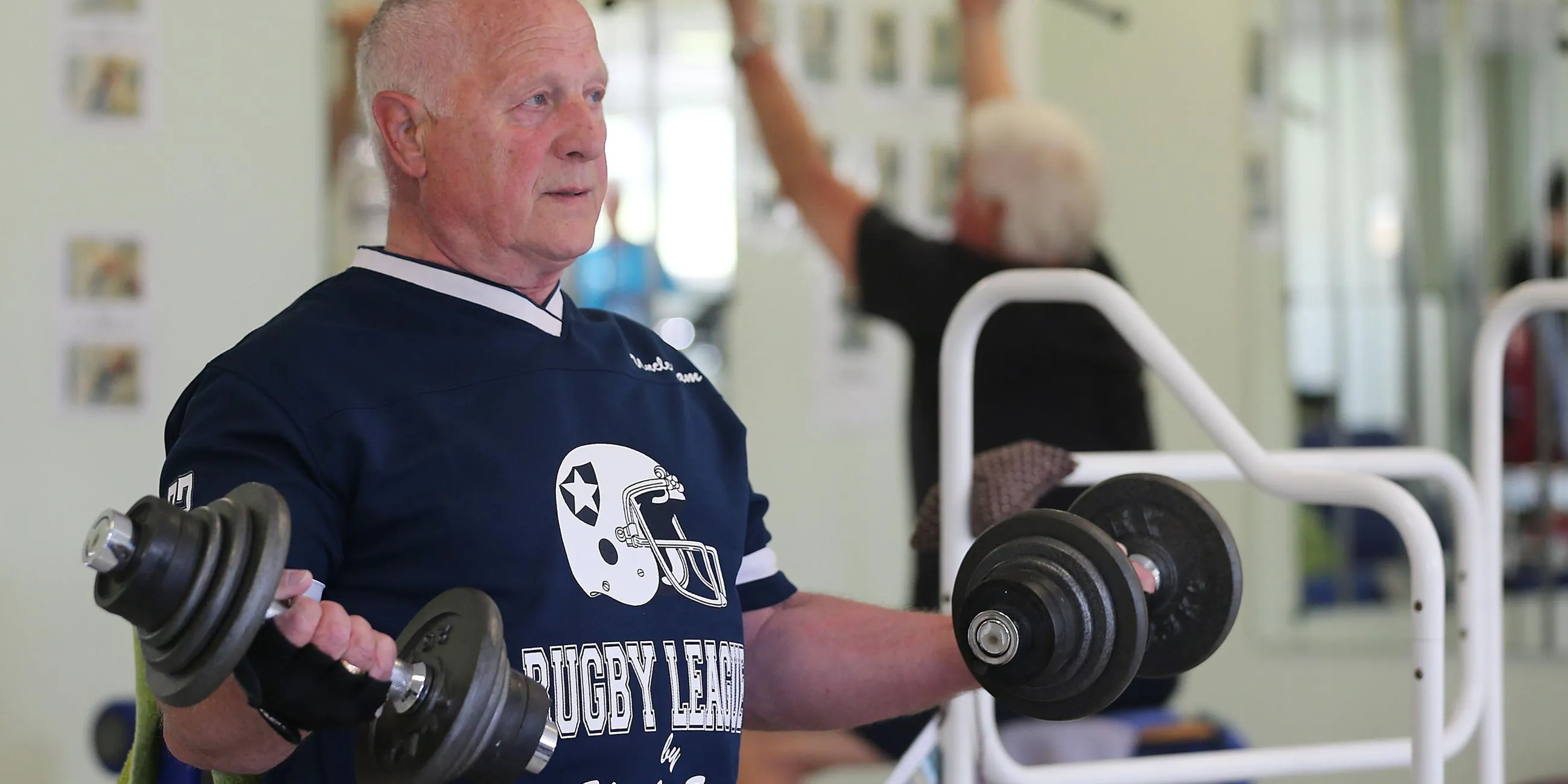 Älterer Herr sitzt in einem Fitnessstudio und macht Hantelübungen.