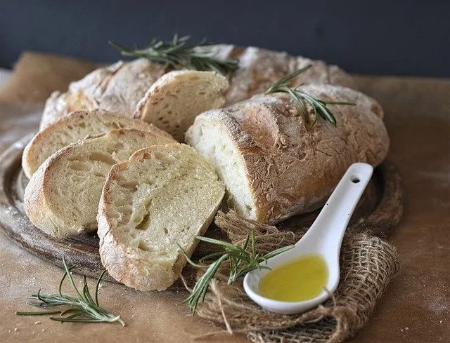 Auf einem rustikalen Holzbrett liegt ein frisch gebackenes Ciabatta, umgeben von kleinen Rosmarinzweigen. Daneben befindet sich ein Löffel mit Olivenöl.