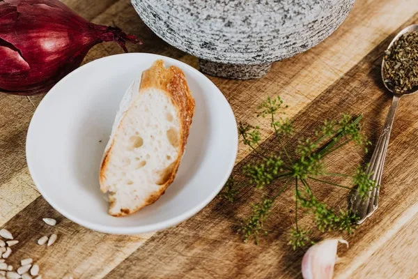 Auf einem rustikalen Holztisch steht ein Mörser, gefüllt mit verschiedenen Gewürzen. Daneben liegt eine Scheibe frisch gebackenes Brot.