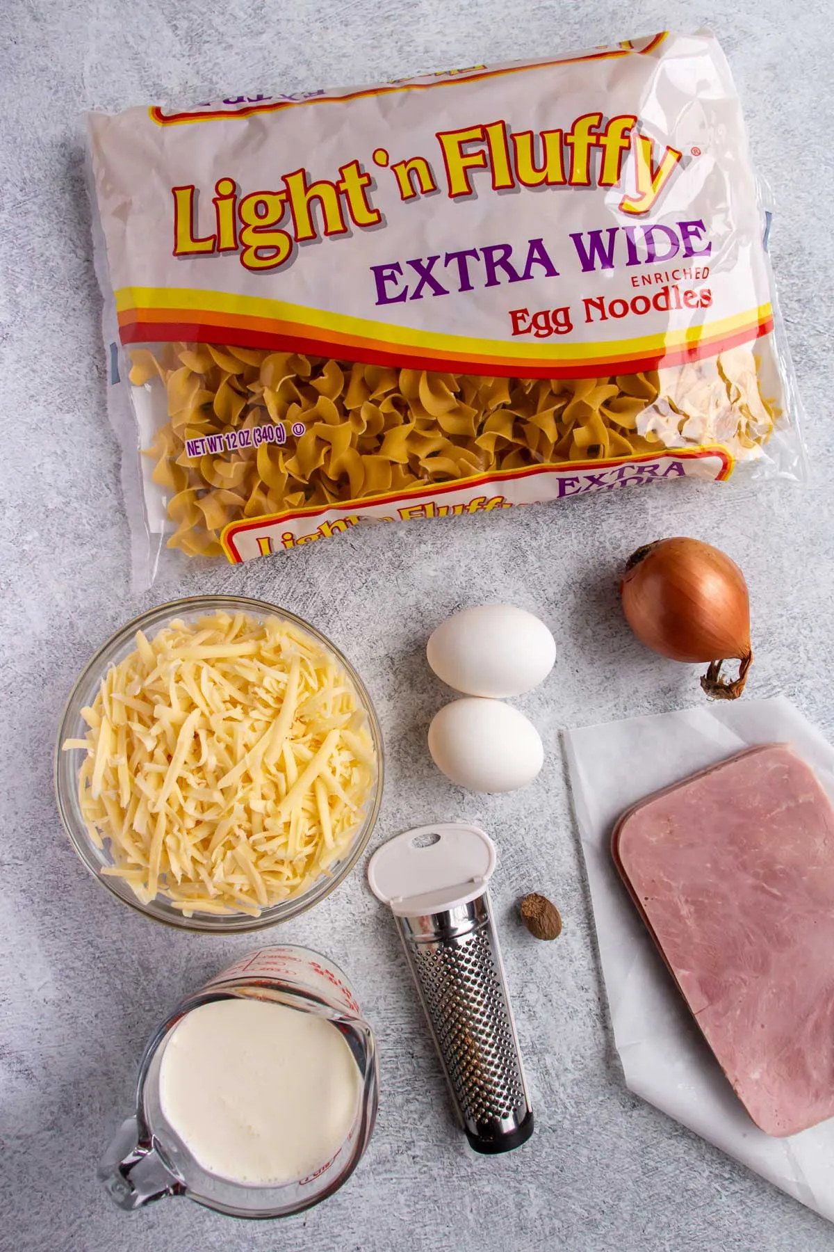 Ausgelegte Zutaten für Schinkennudeln auf grauem Betonhintergrund: Nudeln, Schinken, Käse, Milch, Eier