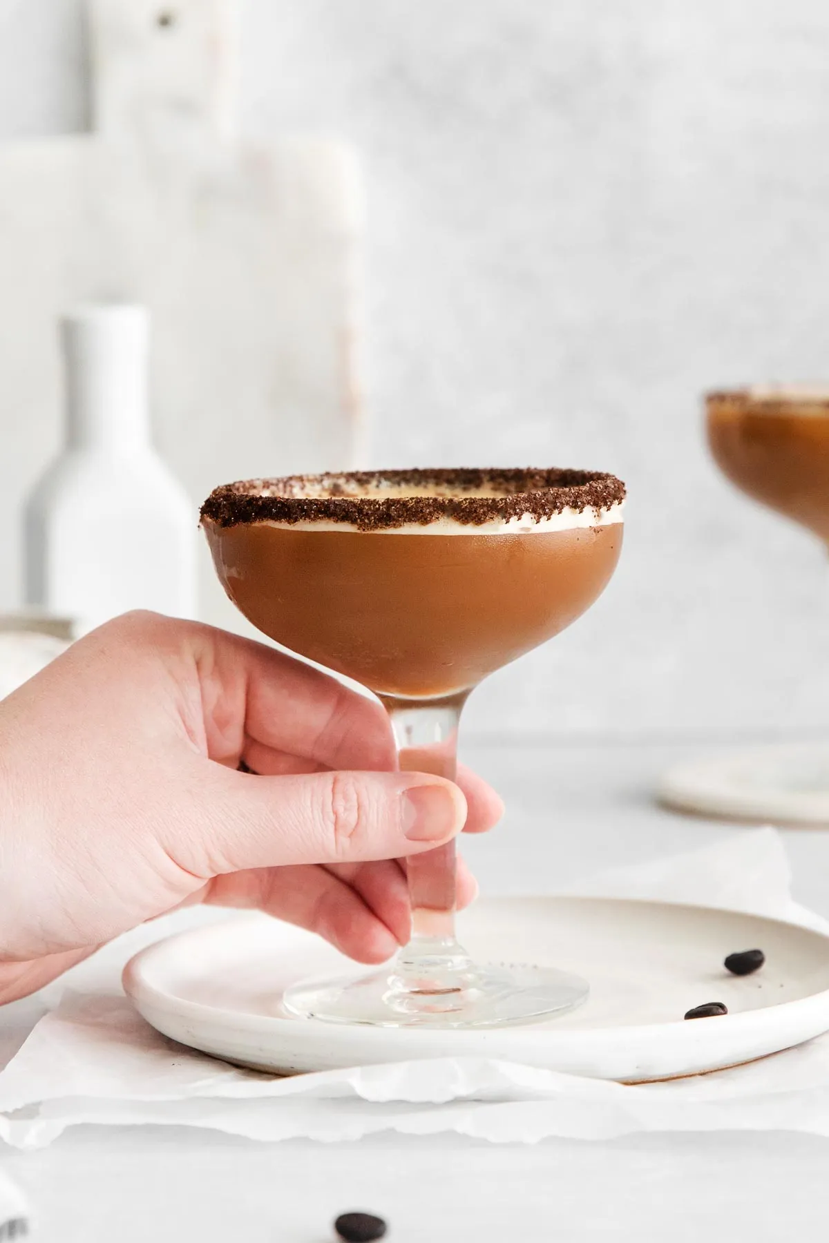 Ein Bild, das eine Hand zeigt, die den Stiel eines Martiniglases voller veganem Espresso Martini hält, dessen Rand mit Kaffeemehl verziert ist