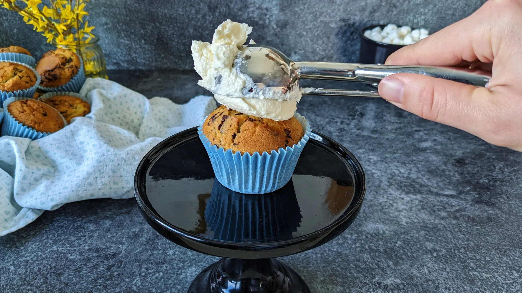 Ein Eisportionierer füllt Muffin-Förmchen mit Teig, daneben ein fertig dekorierter Schaf-Muffin
