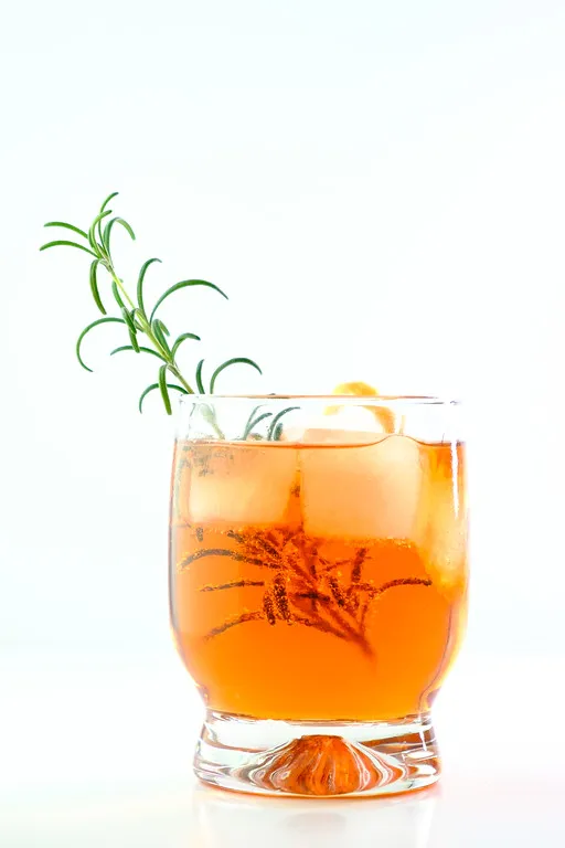 Ein Glas mit einem eleganten orangefarbenen Cocktail, verziert mit einem duftenden Rosmarinzweig