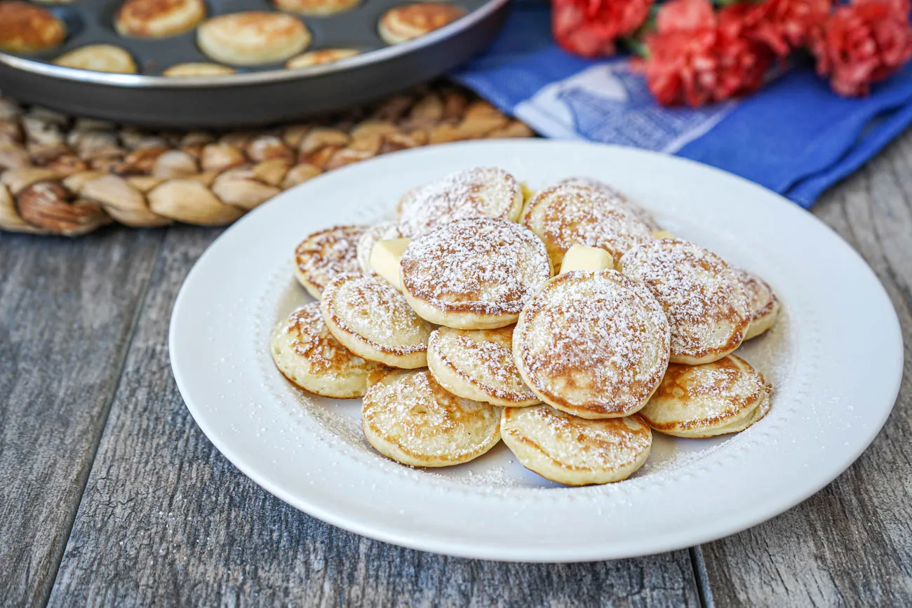 Ein Haufen Poffertjes (Holländische Mini-Pfannkuchen) auf einem weißen Teller, serviert mit Butter und Puderzucker.