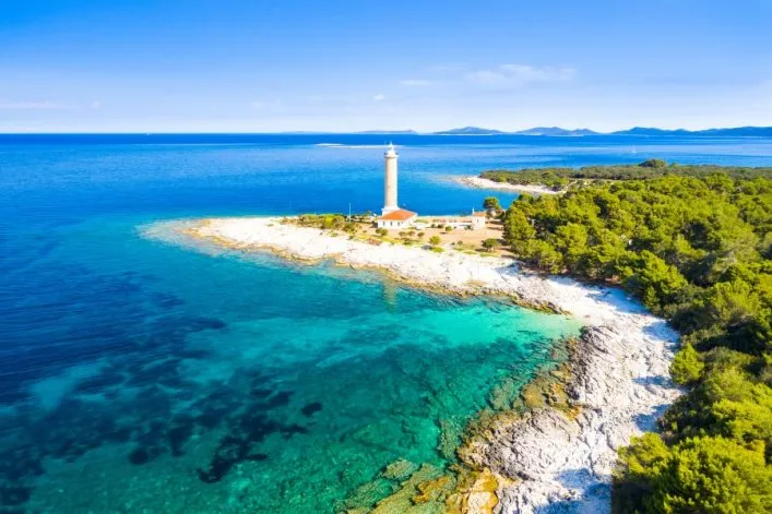 Ein Leuchtturm auf einer felsigen Küste, umgeben von smaragdgrünem Wasser und Pinienwäldern. Der Himmel ist klar blau. Im Hintergrund erstreckt sich das Meer mit fernen Hügeln am Horizont.