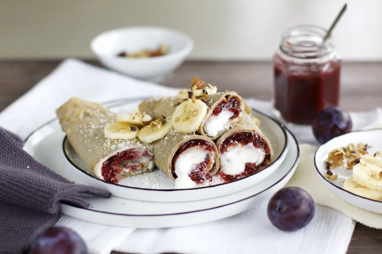 Ein Stapel goldbrauner Buchweizen-Crêpes, serviert mit frischem Pflaumenmus und Nüssen auf einem Holztisch.
