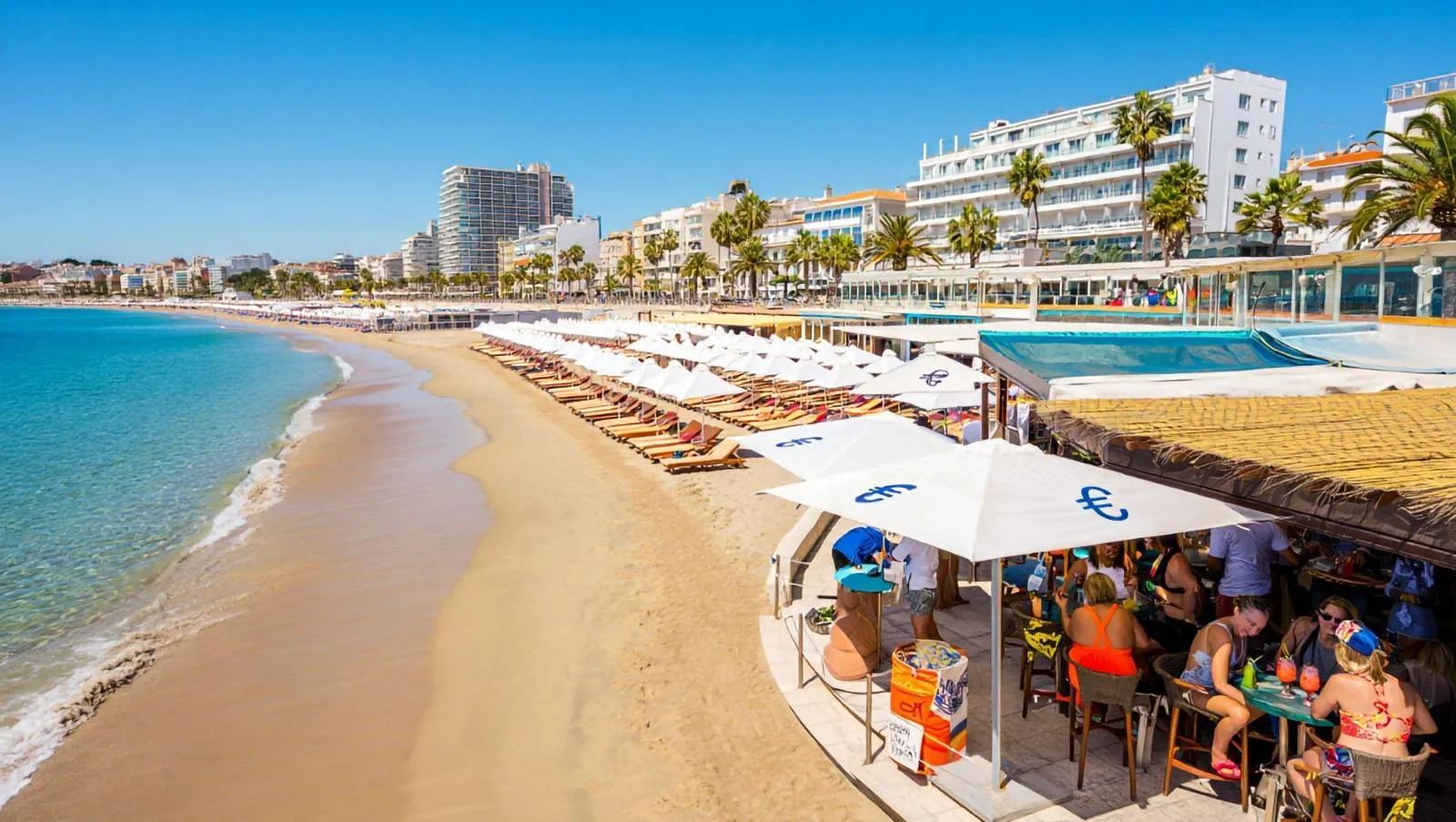 Ein Strand mit mehreren Sonnenliegen und einer Bar, an der Menschen sitzen.