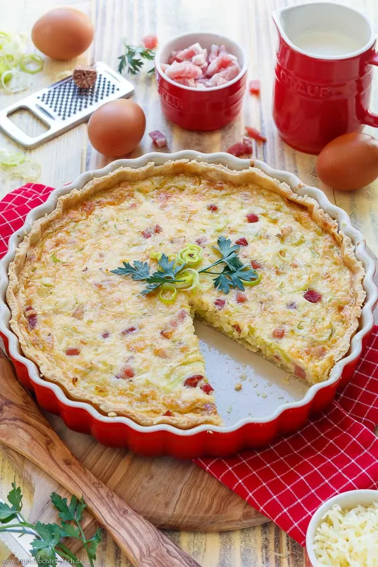 Ein Stück Quiche Lorraine wird aus einer runden Form gehoben, mit goldbrauner Kruste