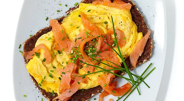Ein Teller mit cremigem Räucherlachs-Rührei, garniert mit frischem Schnittlauch und einer Scheibe Vollkornbrot.