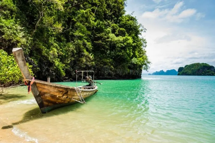 Ein traditionelles Holzboot liegt am sandigen Ufer eines türkisfarbenen Meeres. Im Hintergrund ragen grüne, bewaldete Felsen in den Himmel und kleine Inseln sind in der Ferne sichtbar.