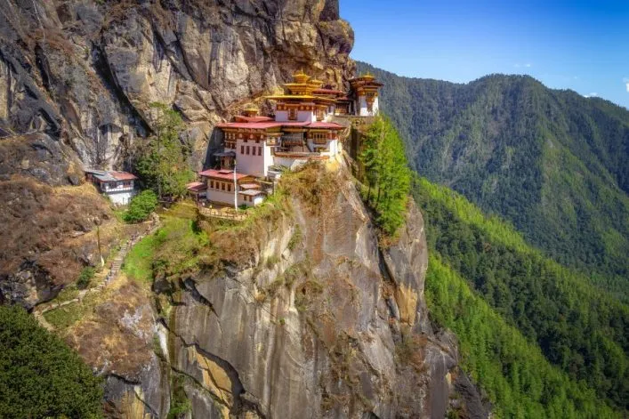 Ein traditionelles Kloster klammert sich an die steile Felswand eines Berges, umgeben von üppigen grünen Bäumen in Bhutan.