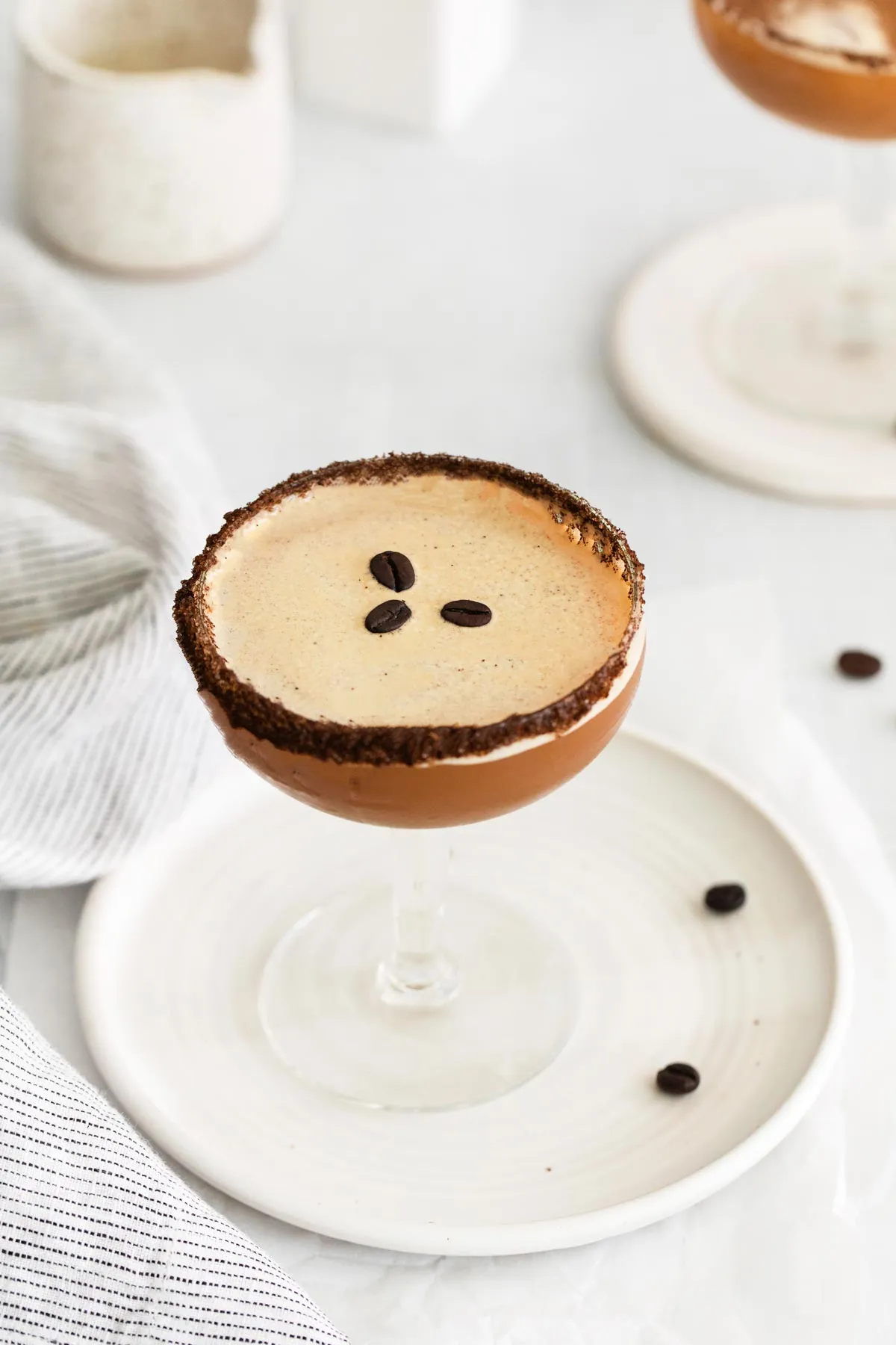 Ein von oben aufgenommenes Bild eines veganen Espresso Martinis in einem Martiniglas auf einem weißen Teller, garniert mit drei gerösteten Kaffeebohnen