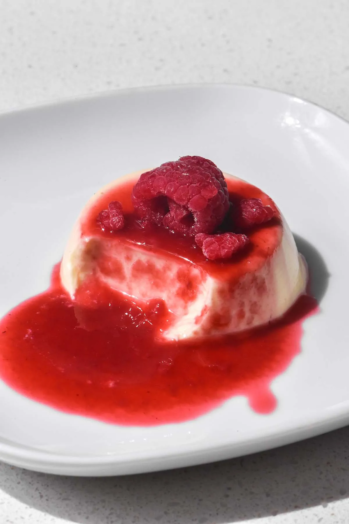 Eine Nahaufnahme einer Panna Cotta auf einem weißen Teller, garniert mit Himbeersauce und ganzen Himbeeren