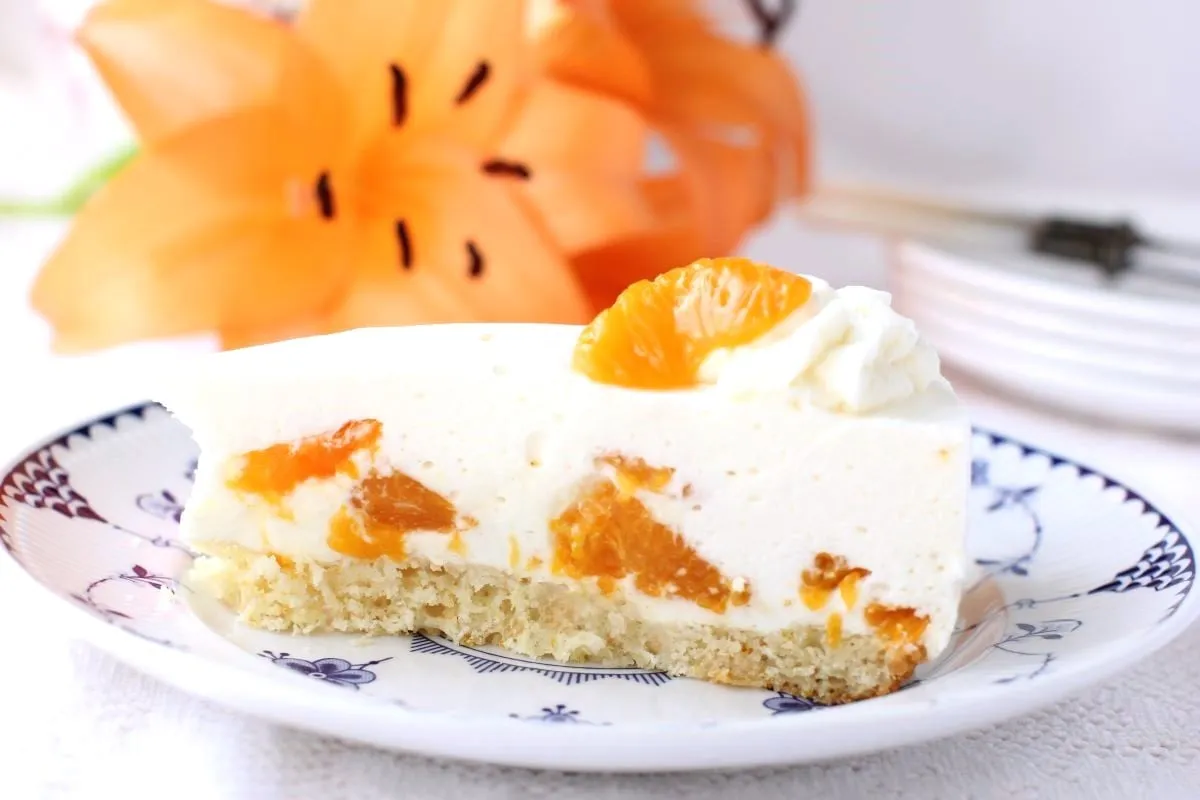Eine Scheibe Mandarinen-Sahnetorte mit einer orangefarbenen Lilienblüte im Hintergrund.