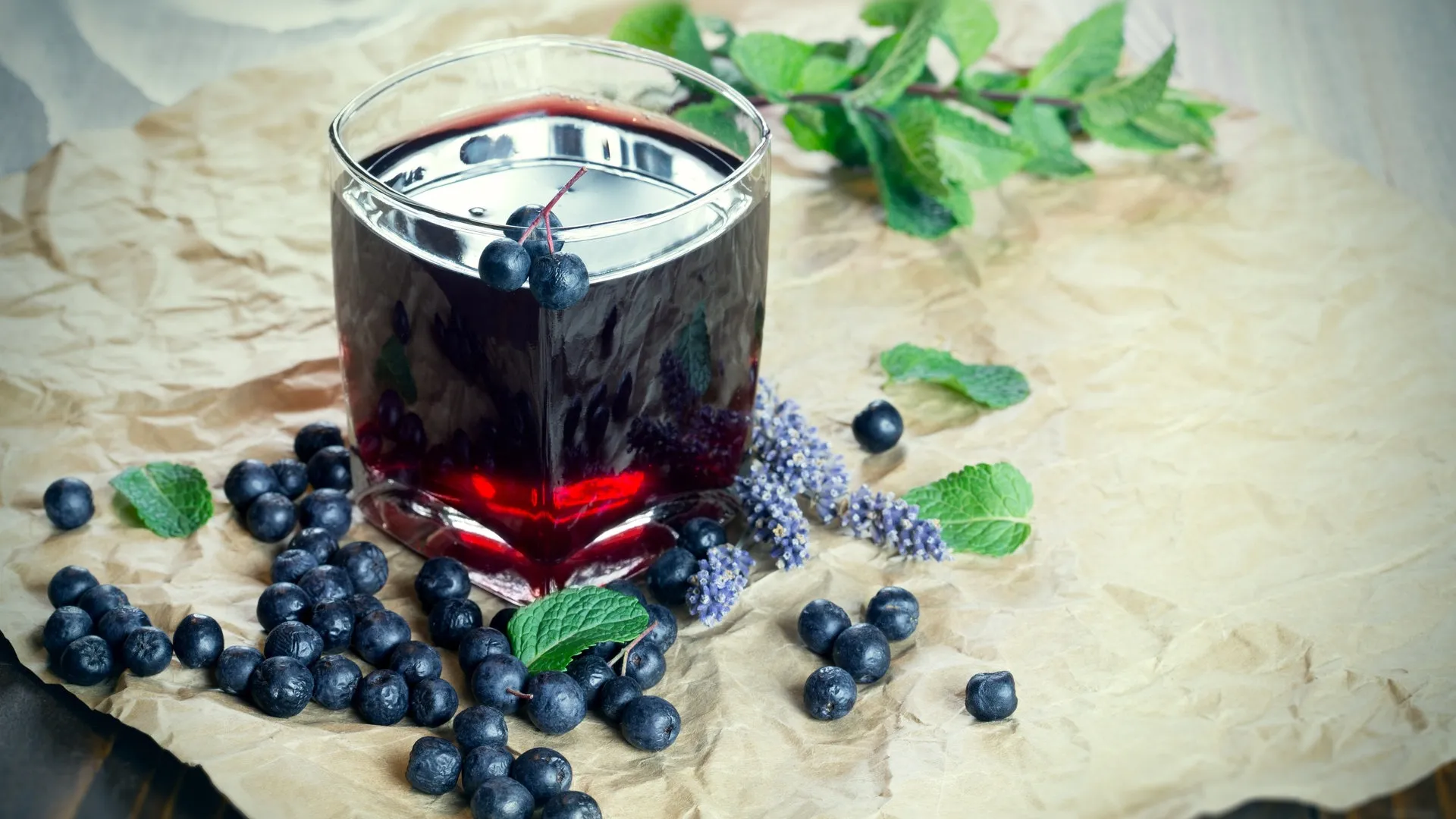 Glas mit dunkelrotem Saft, umgeben von Aronia-Beeren. Aronia soll gut für Immunsystem, Cholesterinwerte und den Blutdruck sein und sogar Krebszellen hemmen. Was ist dran? Kann man die Beeren roh essen?