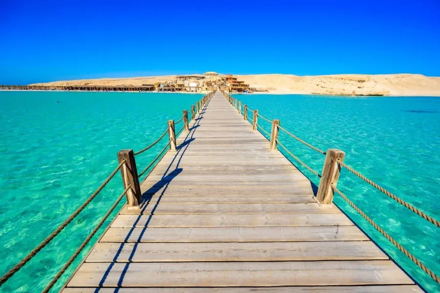 Holzsteg an der Orange Bay in Hurghada: kristallklares Wasser und weißer Sandstrand mit Insel Giftun im Hintergrund.