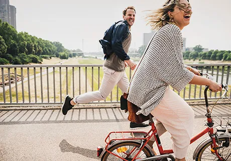 Junge Frau fährt lachend Fahrrad auf der Brücke, begleitet von einem Mann zu Fuß.