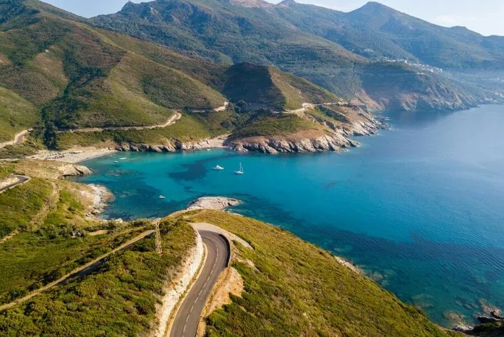 Kurvige Küstenstraße auf Korsika, umgeben von grünen Hügeln, führt zu einer türkisfarbenen Bucht