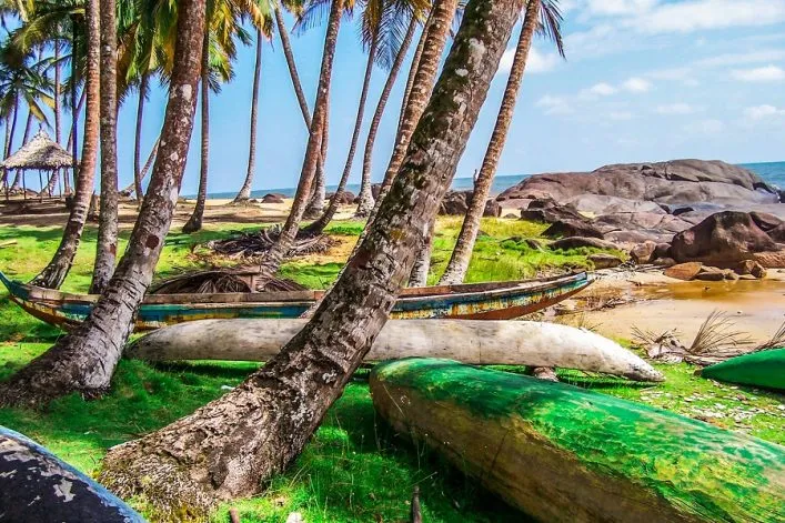 Liberia: Palmengesäumte Küste mit umgedrehten Booten am Sandstrand unter blauem Himmel. Im Hintergrund eine Hütte.
