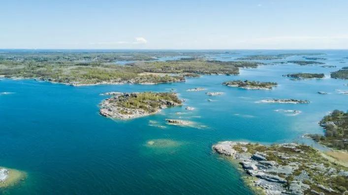 Luftaufnahme der Åland-Inseln; zahlreiche kleine, grüne Inseln sind in türkisblauem Wasser verteilt. Am Horizont erstreckt sich das Festland, das von einem klaren, blauen Himmel umrahmt wird.