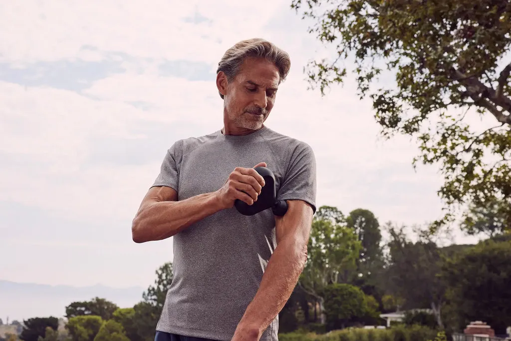 Mann steht in einem Garten und massiert seinen linken Oberarm mit einer Theragun mini.