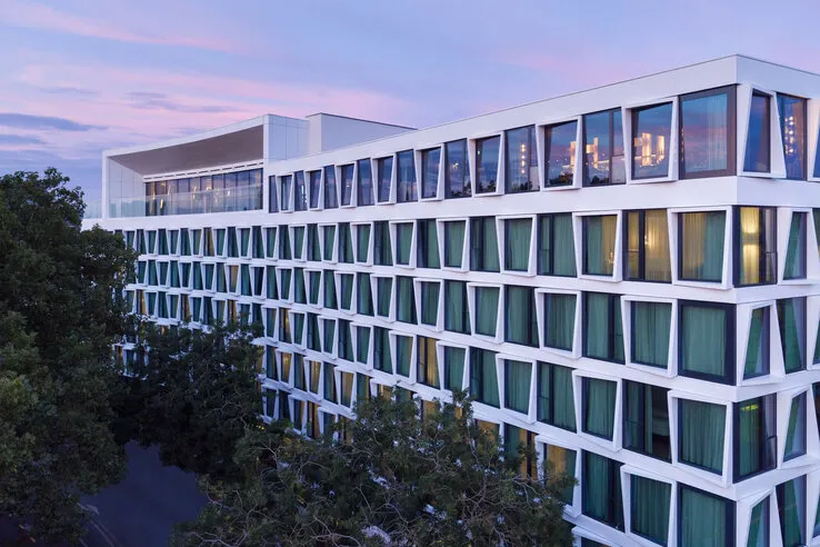 Modernes Hotelgebäude mit großen Fenstern, grünen Vorhängen, umgeben von Bäumen, bei Sonnenuntergang.