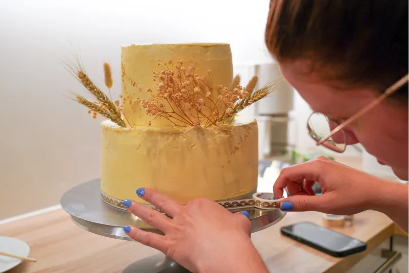 Nahaufnahme der letzten Handgriffe bei der Dekoration einer Hochzeitstorte mit Trockenblumen und Ähren, kurz vor dem großen Fest.