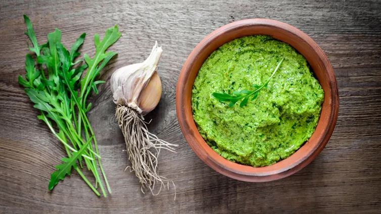Rucola-Pesto in einem kleinen Tontopf, daneben frischer Rucola und Knoblauch