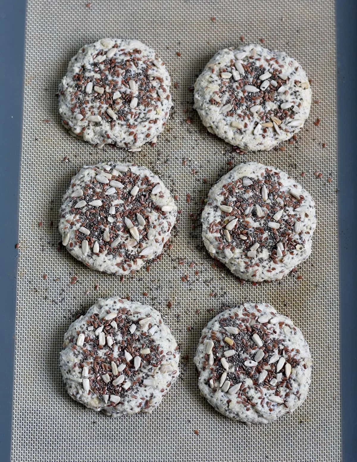 Sechs glutenfreie Brötchen mit Körnern liegen auf einer Backunterlage, bereit zum Backen