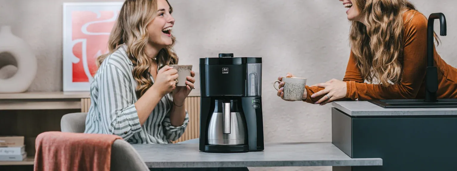 Zwei Frauen genießen frisch gebrühten Kaffee aus einer modernen Melitta LOOK Therm Filterkaffeemaschine.