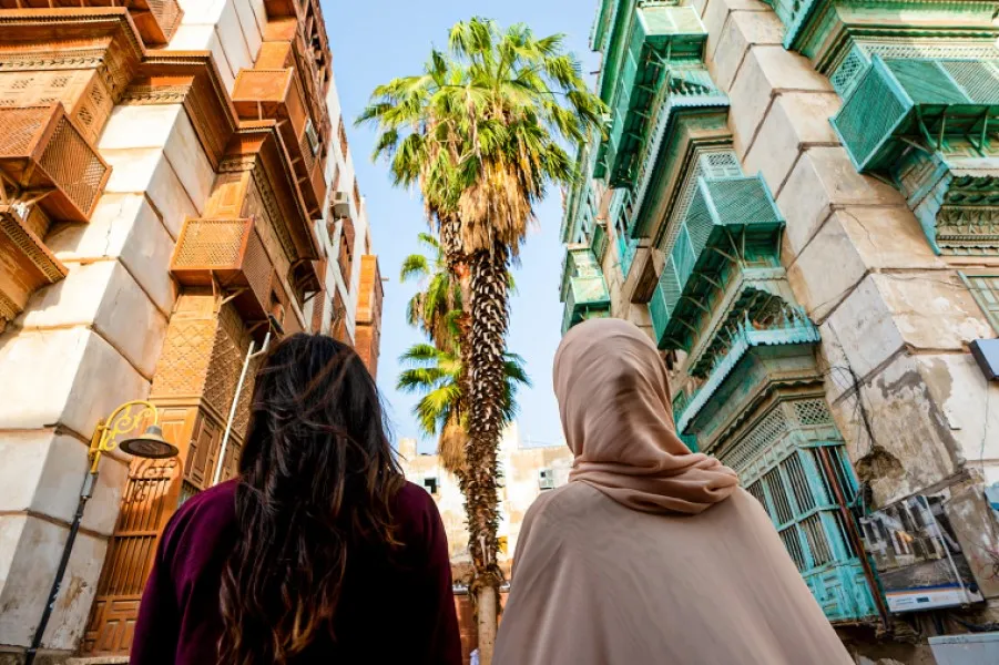 Zwei Frauen spazieren durch eine Gasse der historischen Altstadt Al-Balad in Jeddah, umgeben von bunten Häuserfassaden und Palmen.