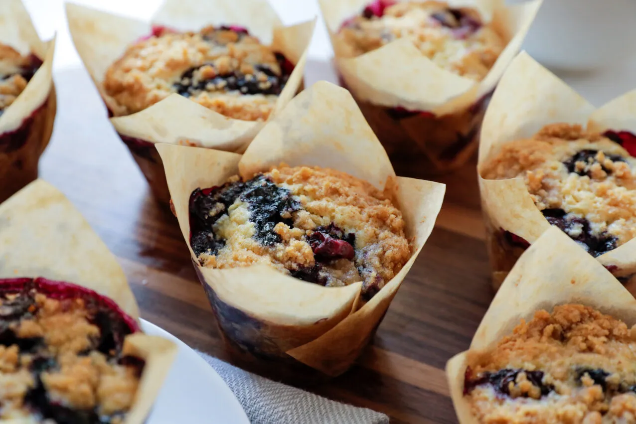 Zwei goldbraune Blaubeer-Muffins mit Streuseldecke, die in kleinen Kupferförmchen gebacken wurden, auf einer dunklen Oberfläche