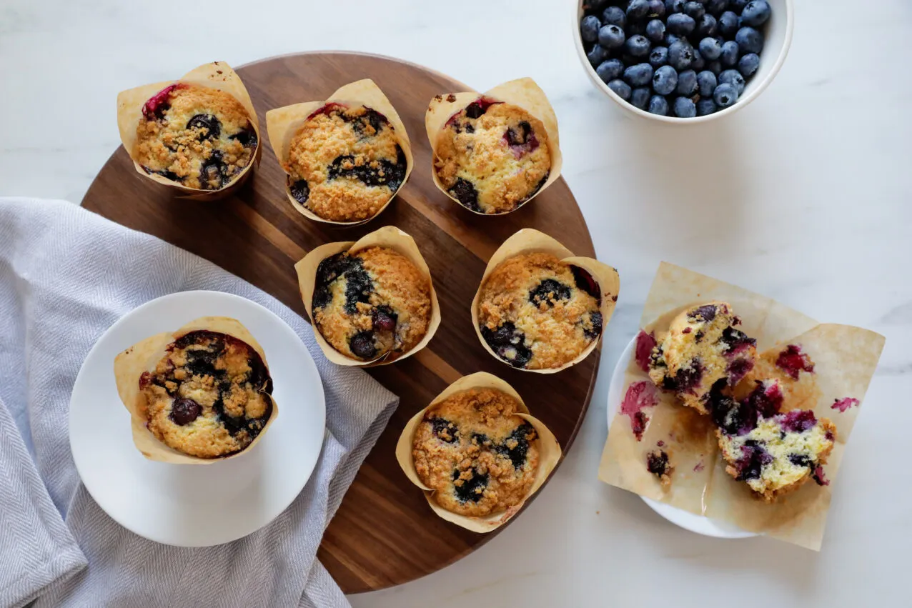 Zwei große, goldbraune Blaubeer-Muffins mit Streuseldecke, die in Tulpencups auf einem Holztisch präsentiert werden