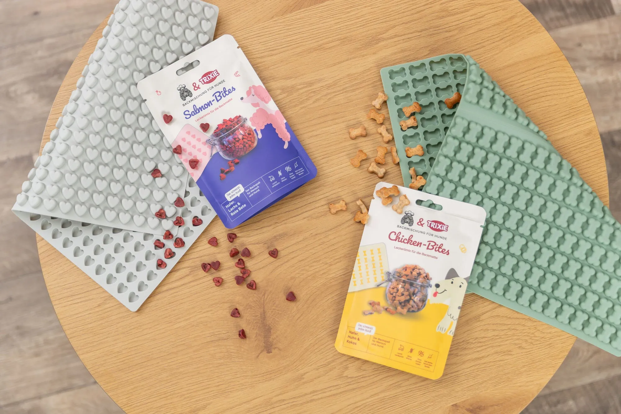 Zwei TRIXIE Backmischungen für Hunde – Salmon Bites und Chicken Bites – liegen auf einem runden Holztisch, umgeben von passenden Silikon-Backmatten in Herz- und Knochenform. Neben den Verpackungen sind gebackene Leckerlis zu sehen – perfekt für gesunde, selbstgemachte Hundesnacks.